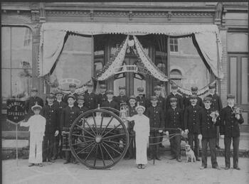 Two rows of uniformed men around a horse drawn but horse-less fire engine